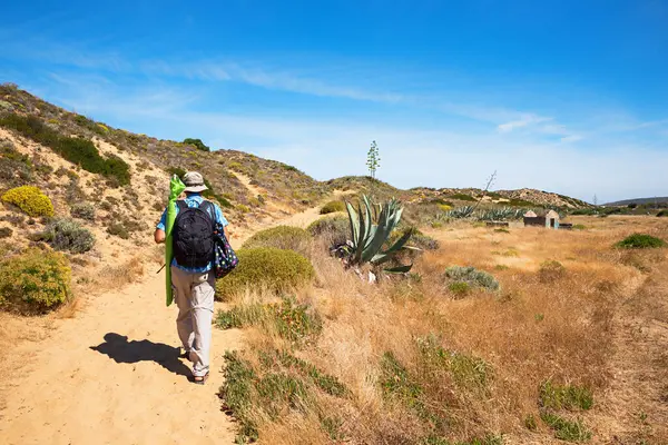 Sırt çantalı ve plaj ekipmanlı bir adam kumlu bir kumul yolu boyunca yürüyordu. Ağaç bitkileri ve Akdeniz çiçekleriyle dolu bir manzara. Portekiz, Costa Vicentina 'da yaz tatili. Boşluğu kopyalayan mavi gökyüzü. 