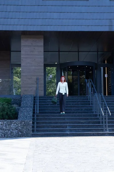 Şık kıyafetler içinde güzel bir kızıl kız ve iş binasının yanında bir çanta.