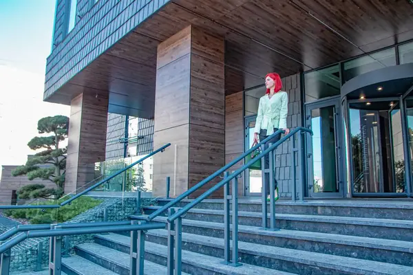 Şık giyinmiş güzel kızıl saçlı bir kız ve el çantasıyla bir iş binasından çıkıyor.