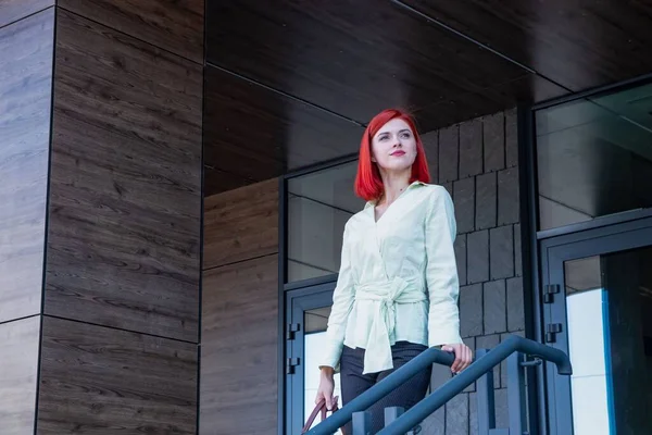 Şık giyinmiş güzel kızıl saçlı bir kız ve el çantasıyla bir iş binasından çıkıyor.