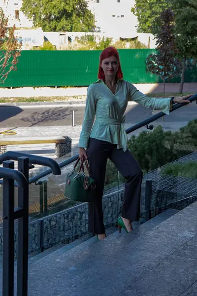 Kızıl saçlı genç bir kadın modern bir iş merkezinin merdivenlerinde duruyor. Büyük cam kapılı, şık ve zarif giyinmiş..