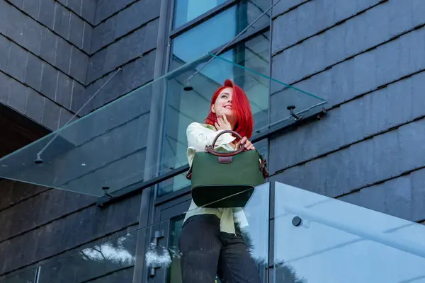 Parlak kızıl saçlı genç bir kadın modern bir binanın yanında elinde şık yeşil bir çanta duruyor..