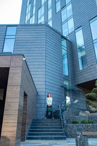 Geniş pencereli ve koyu taş kaplamalı modern bir bina. Ön girişte, kırmızı saçlı bir kadın merdivenlerde dikiliyor. Mimari, modern bir tasarım ile kısıtlanmış ve minimalist görünüyor.