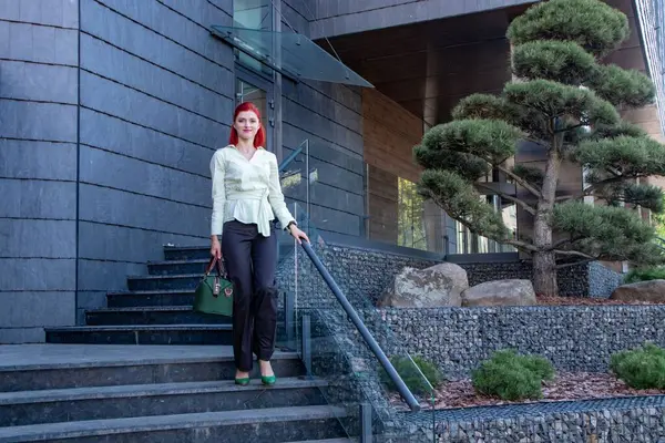 Parlak kızıl saçlı genç bir kadın modern bir binanın yanında elinde şık yeşil bir çanta duruyor..
