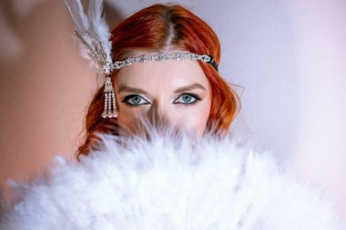 A woman with red hair holding a white fluffy scarf covering half of her face. Elegant retro clothing accessorized with a statement pearl bracelet and feather and sequin headdress. clipart