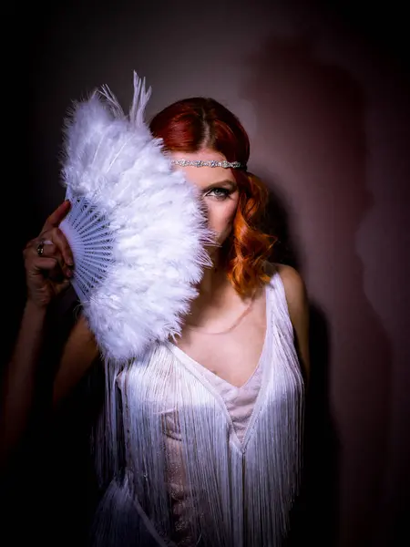 Kızıl saçlı bir kadın eski moda pozlar veriyor. O, püsküllü beyaz bir elbise giyiyor ve şık bir taç başını süslüyor. Yüzünün bir kısmı beyaz tüylü bir vantilatörle kaplanmış. Gizemli bir hava katıyor. Arka plan yumuşak gölgelerle karanlık..