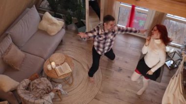 Genç bir çift büyük pencereli kiralık bir ahşap evde aktif olarak dans ediyor ve şarkı söylüyor. Yukarıdan bak. Küçük bir evde dinlen.