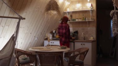 Flanel gömlekli bir kadın bir şişe açar ve aperatiflerle yuvarlak bir masanın yanında dururken bardağa şarap döker. Konforlu mutfak İskandinav ya da eko tarzında dekore edilmiştir..