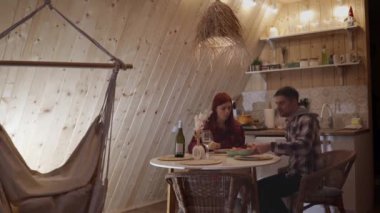 Genç bir çift, ahşap duvarları olan rahat bir mutfakta yuvarlak bir masada oturuyor. Şarap içip karides yiyorlar. İçi, minimalist dekorla süslenmiş..