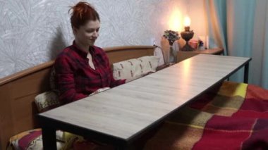 Kırmızı gömlekli bir kadın, üzerinde suşi, bardakta içki ve mumların servis edildiği bir masanın önündeki yatağa oturur. Eve teslim konsepti