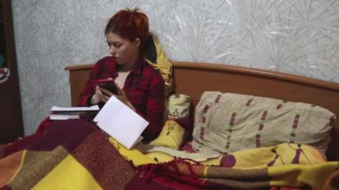 Güzel bir kadın yatakta, battaniyeye sarılmış ve çalışıyor. O kırmızı bir gömlek giyiyor, bir akıllı telefon tutuyor ve aktif olarak onu kullanıyor, yanında bir defter ve bir kitap yatıyor..