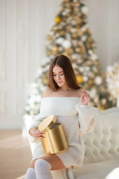 クリスマスを祝う幸せな若い美しい女性 白いセーターの女の子はクリスマスツリーの背景に贈り物を開きます クリスマスのコンセプト ギフトボックスと笑顔の女性 — ストック写真