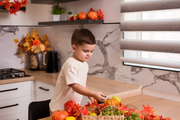 Mutfak masasında meyveler, sonbahar yaprakları ve balkabağıyla otururken çok tatlı gülümsüyor. Sonbahar dekore edilmiş mutfak. Tatil kavramı