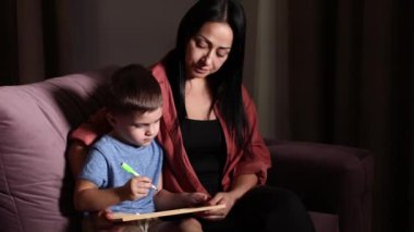 Kapalı alanda renkli kalemler ve keçeli kalemlerle çizim yapan sevimli bir çocuk ve genç bir anne. Güzel anne resme yardım ediyor. Güzel anne ve çocuk evde birlikte vakit geçiriyorlar.