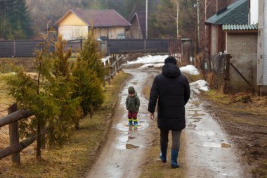 Baba ve oğul temiz havada, su birikintilerinde lastik botlarla yağmurdan sonra çamurda yürüyorlar. Küçük çocuk bir adamın elini tutuyor..