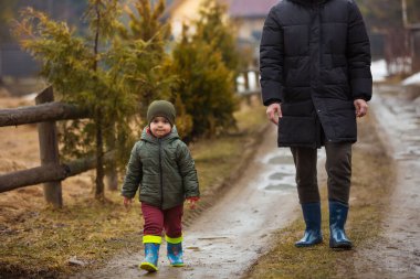 Baba ve oğul temiz havada, su birikintilerinde lastik botlarla yağmurdan sonra çamurda yürüyorlar. Küçük çocuk bir adamın elini tutuyor..