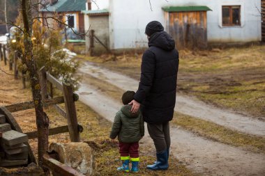 Baba ve oğul temiz havada, su birikintilerinde lastik botlarla yağmurdan sonra çamurda yürüyorlar. Küçük çocuk bir adamın elini tutuyor..
