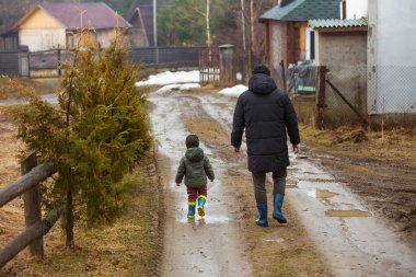 Baba ve oğul temiz havada, su birikintilerinde lastik botlarla yağmurdan sonra çamurda yürüyorlar. Küçük çocuk bir adamın elini tutuyor..