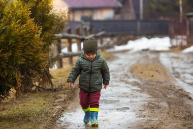 Koruyucu lastik botları ve çamur birikintisinde zıplayan yağmurluk giymiş küçük bir çocuk. Mutlu çocuk yağmur üstüne su birikintisinde oynarken eğleniyor.