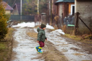 Koruyucu lastik botları ve çamur birikintisinde zıplayan yağmurluk giymiş küçük bir çocuk. Mutlu çocuk yağmur üstüne su birikintisinde oynarken eğleniyor.