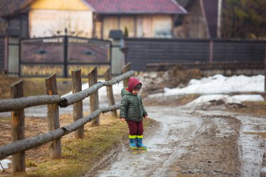 Koruyucu lastik botları ve çamur birikintisinde zıplayan yağmurluk giymiş küçük bir çocuk. Mutlu çocuk yağmur üstüne su birikintisinde oynarken eğleniyor.