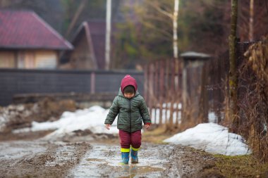 Koruyucu lastik botları ve çamur birikintisinde zıplayan yağmurluk giymiş küçük bir çocuk. Mutlu çocuk yağmur üstüne su birikintisinde oynarken eğleniyor.