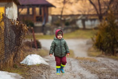 Koruyucu lastik botları ve çamur birikintisinde zıplayan yağmurluk giymiş küçük bir çocuk. Mutlu çocuk yağmur üstüne su birikintisinde oynarken eğleniyor.
