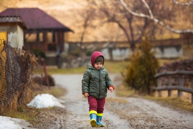 Koruyucu lastik botları ve çamur birikintisinde zıplayan yağmurluk giymiş küçük bir çocuk. Mutlu çocuk yağmur üstüne su birikintisinde oynarken eğleniyor.