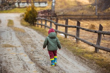 Koruyucu lastik botları ve çamur birikintisinde zıplayan yağmurluk giymiş küçük bir çocuk. Mutlu çocuk yağmur üstüne su birikintisinde oynarken eğleniyor.