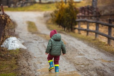Koruyucu lastik botları ve çamur birikintisinde zıplayan yağmurluk giymiş küçük bir çocuk. Mutlu çocuk yağmur üstüne su birikintisinde oynarken eğleniyor.