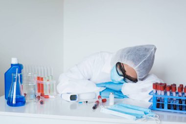Stressed female doctor sitting tired at his desk. Mid adult female doctor working long hours in protective clothes. Overworked doctor in his office needs a break. Focus on hand