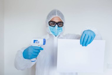 Female doctor with protective workwear holding empty cardboard while standing at clinic and looking at camera. Copy space.