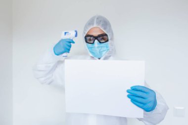 Female doctor with protective workwear holding empty cardboard while standing at clinic and looking at camera. Copy space.