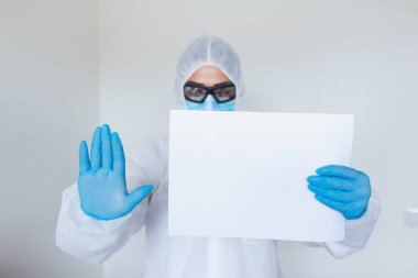Female doctor with protective workwear holding empty cardboard while standing at clinic and looking at camera. Copy space.