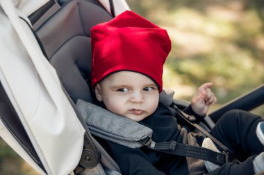 Sevimli kırmızı şapkalı çekici bir çocuk dışarıda bir bebek arabasında mutlu bir şekilde otururken ışıl ışıl gülümsüyor.