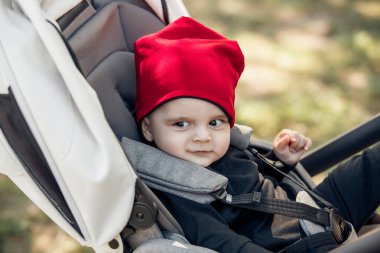 Sevimli kırmızı şapkalı çekici bir çocuk dışarıda bir bebek arabasında mutlu bir şekilde otururken ışıl ışıl gülümsüyor.