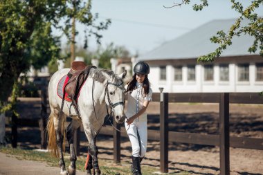 Manzaralı bir binicilik ortamında, genç bir binici güzel beyaz bir atın yanında yürüyor, anın tadını çıkarıyor.