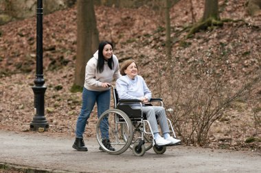 Yürek ısıtan bir sahne, neşeli doğayla dolu sakin bir parkta tekerlekli sandalyeyi iten bir bakıcıyı gösteriyor.
