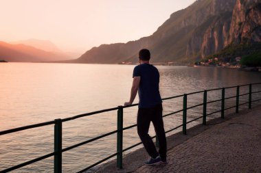 Yalnız bir adam, altın saat boyunca dağlarla çevrili huzurlu bir göle bakarak korkuluklara yaslanıyor..