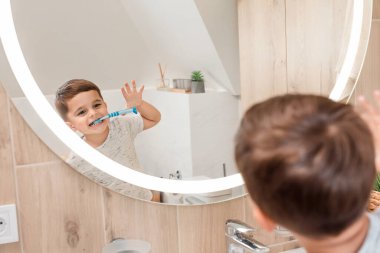 Genç bir çocuk aynada komik bir yüz ifadesi takınırken neşeyle dişlerini fırçalar. Yansıması, parlak ve modern banyoyla çevrili günlük rutinin enerjisini ve eğlencesini yakalıyor..