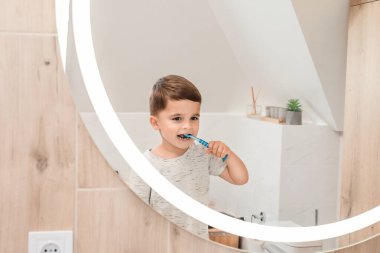 Genç bir çocuk aynada komik bir yüz ifadesi takınırken neşeyle dişlerini fırçalar. Yansıması, parlak ve modern banyoyla çevrili günlük rutinin enerjisini ve eğlencesini yakalıyor..