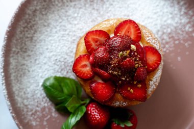 Taze çilek, pudra şekeri ve fesleğen yapraklarıyla kaplı lezzetli bir mini pasta, yumuşak pembe bir tabakta güzelce servis edilir..