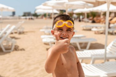 Sarı yüzme gözlüğü takan gülümseyen genç bir çocuk güneşli bir sahilde başparmağını kaldırıyor. Şemsiyeler ve şezlonglar arkada bulanıklaşıyor. Güven ve eğlence dolu neşeli bir yaz anıydı..