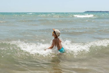 Genç Çocuk, kasketli ve yüzme şortlu küçük bir çocuk kumsaldaki dalgalara bakarken kollarını genişçe uzatıyor. Deniz bacaklarının etrafında sıçrıyor, kaygısız, neşeli bir yaz yaz anı yakalıyor..