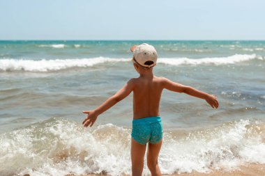Genç Çocuk, kasketli ve yüzme şortlu küçük bir çocuk kumsaldaki dalgalara bakarken kollarını genişçe uzatıyor. Deniz bacaklarının etrafında sıçrıyor, kaygısız, neşeli bir yaz yaz anı yakalıyor..
