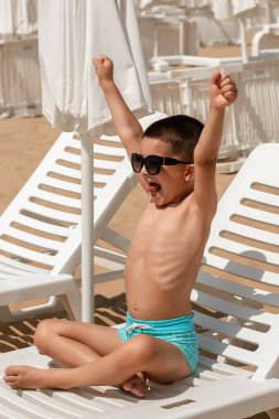 Mavi yüzme şortu ve güneş gözlüğü takmış heyecanlı genç bir çocuk beyaz bir plaj sandalyesinde bağdaş kurarak otururken kollarını havaya kaldırıyor. Güneşli, kumlu plaj arka planda aynı şezlonglar ve şemsiyelerle kaplı..
