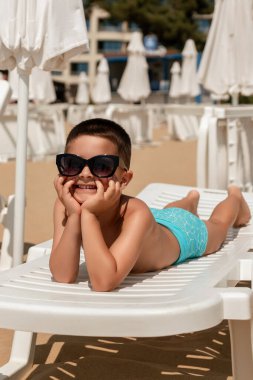 Güneş gözlüklü neşeli bir çocuk beyaz bir plaj sandalyesinde uzanır, kumlu bir sahilde güneşlenir ve etrafında boş sandalyeler vardır..