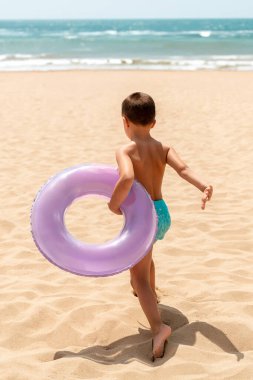 Mor yüzme yüzüğü tutan genç bir çocuk, parlak ve güneşli bir plaj gününde denize doğru yalınayak yürüyor. Saf bir yaz özgürlüğü anı.
