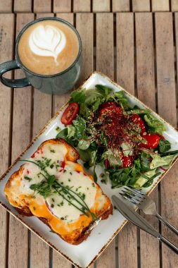Eritilmiş peynirli tost ve taze salatayla servis edilen otlar ve ahşap bir masada bir fincan latte..