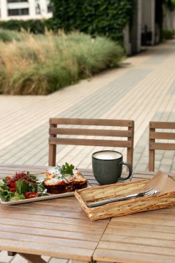 Eritilmiş peynirli tost ve taze salatayla servis edilen otlar ve ahşap bir masada bir fincan latte..